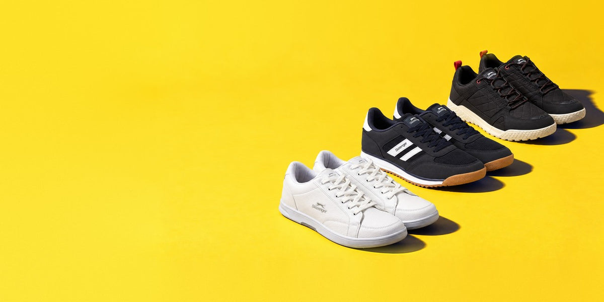 Three pairs of Slazenger sneakers in white, navy and black lined up on a yellow background